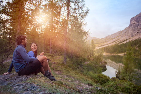Smiling happy man and woman couple enjoy lake panorama view with sun flare light. Group of friends people summer adventure journey in mountain nature outdoors. Travel exploring Alps, Dolomites, Italy.