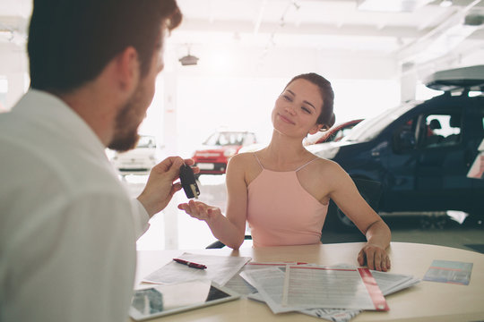 Friendly Car Salesman Talking To A Young Woman And Showing A New Car Inside Showroom Signing Of Contract.