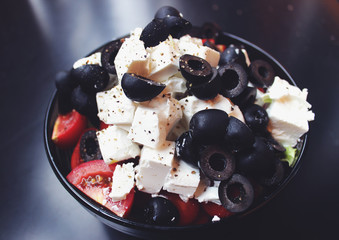 Greek salad with sirtaki cheese and black olives and a red tomato. On white cheese is ground pepper. Salad in a glass plate is on a dark table.