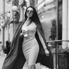 Young beautiful stylish woman with long straight brunette hair walking at the street. Pretty girl in pink elegant dress and coat holding shopping bag and cup of coffee.