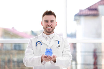 Happy young doctor holding piggy bank on blurred background