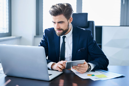 Young confident office worker using tablet and laptop to work in his office