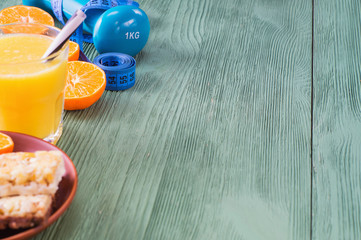 The concept of healthy lifestyle with copy space. Orange juice, fruits. cereal bars, dumbbell and measuring tape on green wooden table. Cereals and fruits - diet and breakfast