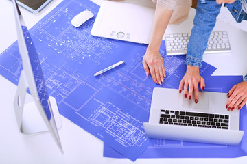 Two young woman standing near desk with instruments, plan and laptop.