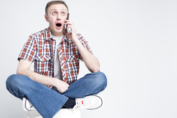 Portrait of Surprised Caucasian Man in Checkered Shirt and Jeans Talking on Cellphone Against White Background. Sitting on White Box.