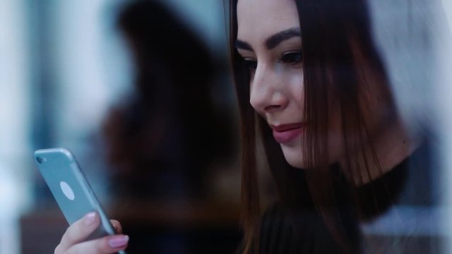 Close up view of a young attractive girl sitting by the window in a cozy caf&eacute; texting the message with her phone, reacting happily on the reply, drinking hot coffee. Modern lifestyle, morning routine.