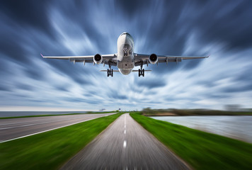 Fototapeta premium Airplane and road with motion blur effect. Landscape with white passenger airplane is flying in the cloudy sky over the asphalt road. Blurred. Passenger airplane is landing. Commercial plane. Aircraft