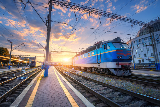 Fototapeta Blue train at the railway station at sunset. Amazing industrial landscape with old locomotive, old buildings, rails and colorful sunset sky with clouds. Train on railroad. Beautiful railway platform