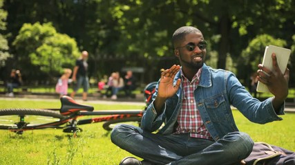 Young african student using tablet in the park while sitting on the grace on background with bikecycle and green tree.