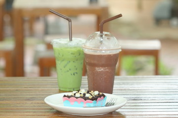 Brownie chocolate cake have marshmallow, almond, chocolate- chip on topping and have fork beside on white dish and soft drink (glass of green tea milk and chocolate milk) on wood table