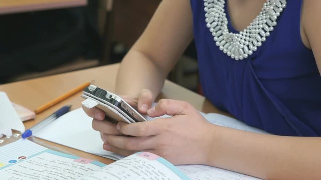 Schoolgirl dressed in blue dress sits at a school desk holds silver smartphone