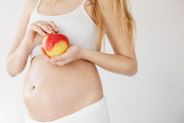 Close up of blonde pregnant woman holding natural apple full of vitamins and iron. Prenatal diet concept. Copy space. High key.