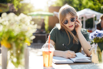 Portrait of a young business woman or student writing her plans in notepad talking on a smartphone wearing glasses during a luch break in park on a sunny summer day.