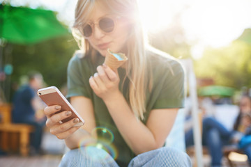 Woman eating icecream using smartphone. Portrait of confused girl with ice cream browsing through social media or messaging her friends enjoying summer in the city park wearing shades. Focus on phone.