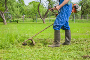 Man wearing overalls and rubber boots mowing green grass with gasoline lawn trimmer. The...