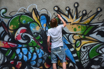 Photo of a young guy in denim shorts and a white shirt. The guy draws on the graffiti wall a drawing with aerosol paints of various colors. The concept of hooliganism and damage to property - obrazy, fototapety, plakaty
