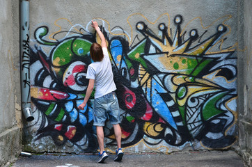 Photo of a young guy in denim shorts and a white shirt. The guy draws on the graffiti wall a drawing with aerosol paints of various colors. The concept of hooliganism and damage to property - obrazy, fototapety, plakaty