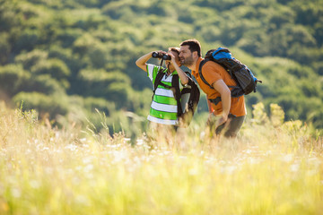 Father and son are hiking in nature in summer. They are using binoculars.
