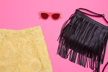 Part of yellow lace shorts, a black bag with fringe and rose-colored glasses. Bright pink background, close-up