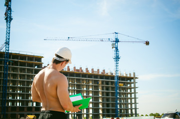 Young sexy male bodybuilder athlete with a bare torso. Manager engineer builder in white protective helmet stands on background of construction of building and construction cranes with documentation