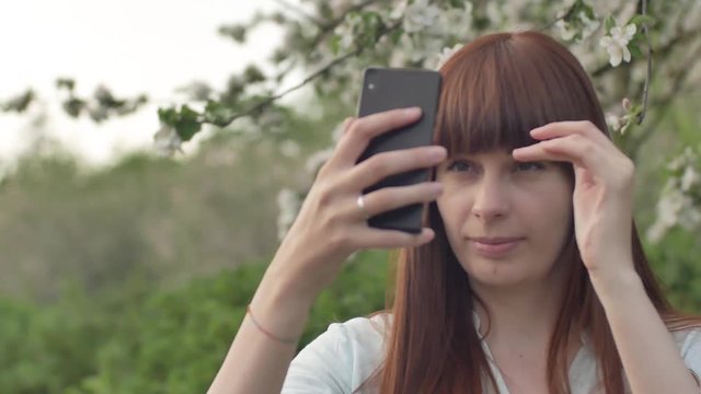 A girl makes selfie in the garden. An attractive red-haired woman smiles making selfi using a mobile phone in a cherry orchard. The concept of using gadgets for a healthy lifestyle. Young sporty happy