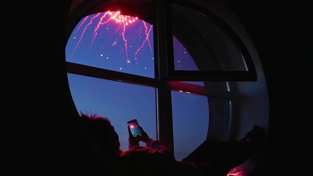 Girl taking fireworks video with a smartphone. Silhouette in a round loft window