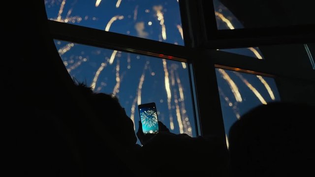Girl taking fireworks video with a smartphone. Silhouette in a round loft window