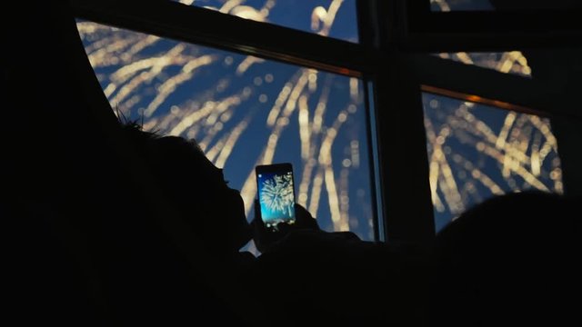 Girl taking fireworks video with a smartphone. Silhouette in a round loft window