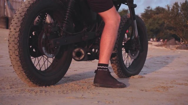 Close up shot of motorcyclist starting to drive his motorbike on the beach during sunset, slow motion
