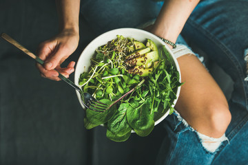 Green vegan breakfast meal in bowl with spinach, arugula, avocado, seeds and sprouts. Girl in jeans holding fork with knees and hands visible, top view, copy space. Clean eating, dieting food concept