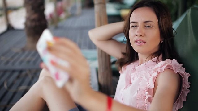 A woman on a lounger drinks a cocktail and takes pictures of herself with a phone