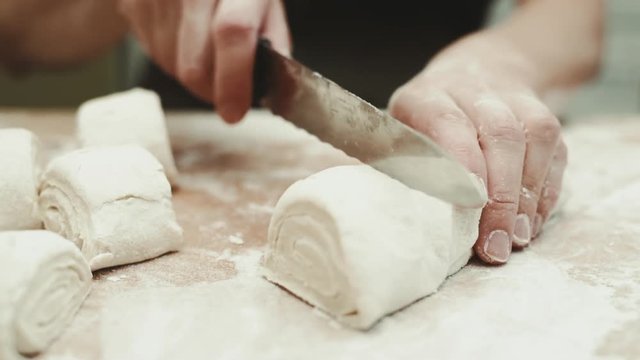 Woman baker cutting raw dough roll into pieces on the table with flour.    