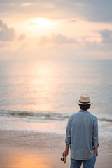 Young Asian man traveler and photographer with jean shirt and hat holding camera and looking beautiful sunset at tropical beach island, background for summer holiday and vacation travel concepts