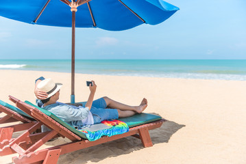 young man lying on wooden beach bench with blue umbrella and take photo of turquoise sea and...