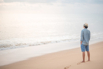 Young Asian man with jean shirt and hat standing on the beach and looking beautiful sunset, vacation time and summer holiday concepts
