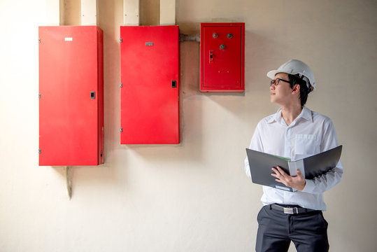 Young Asian Electrical Engineer holding files while wearing a personal protective equipment safety helmet at construction site with red electric control panel. Engineering Maintenance concept.