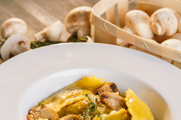 Basket of raw mushrooms and white plate with freshly prepared pasta ravioli and fried champignons cut into slices. The dish is decorated with a sprig of rosemary. Traditional Italian homemade food.