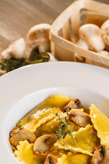 Basket of raw mushrooms and white plate with freshly prepared pasta ravioli and fried champignons cut into slices. The dish is decorated with a sprig of rosemary. Traditional Italian homemade food.