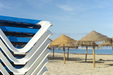 Detail of stacked beach sunbeds with some sunshades and sea in the background