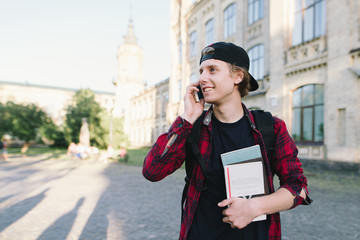 A student in a red shirt, with books in his hands, talks on a mobile phone with his friends on the background of the university and smiles.