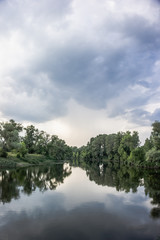 Calm beautiful scenic landscape with blue river, green trees and reflecting in water with cloudy sky. Magical sunset over the river in rural terrain. Natural, wild landscape.