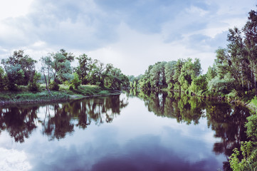 Calm beautiful scenic landscape with blue river, green trees and reflecting in water with cloudy sky. Magical sunset over the river in rural terrain. Natural, wild landscape.