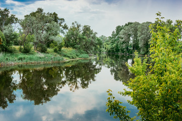 Fototapeta na wymiar Calm beautiful scenic landscape with blue river, green trees and reflecting in water with cloudy sky. Magical sunset over the river in rural terrain. Natural, wild landscape.