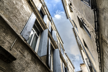Fototapeta na wymiar Strip of blue sky with clouds between two old french close standing buildings windows with weather beaten open shutters