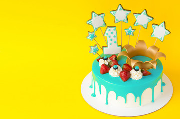 Birthday cake 1 year for little child with cyan glaze, strawberries, cream, blueberry, gold crown, gingerbreads cookies in the form of star, number one. Yellow background. Invitation or greeting card.