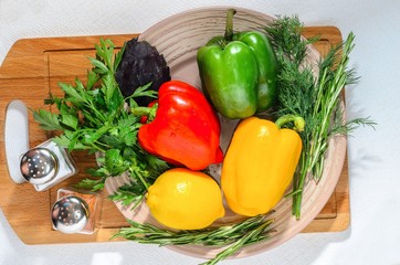 Fresh bright vegetables: Bulgarian pepper, herbs, lemon, on a ceramic plate, on a wooden cutting board