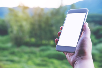 Mockup image of a hand holding and showing white smart phone with blank screen at outdoor and green nature background