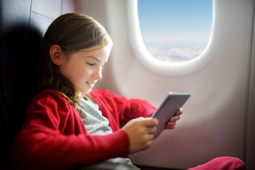 Adorable little girl traveling by an airplane. Child sitting by aircraft window and using a digital tablet during the flight.