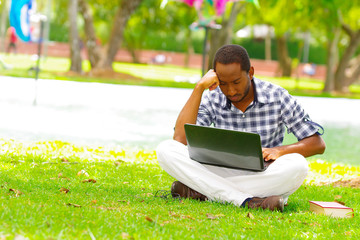Young black man sitting down on green grass and working in his computer in the city of Quito Ecuador