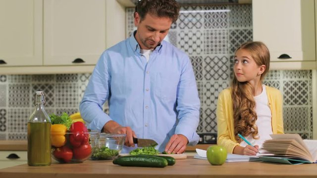 Caucasian family in the kitchen, father cutting the lettuce for salad while girl doing homework.
